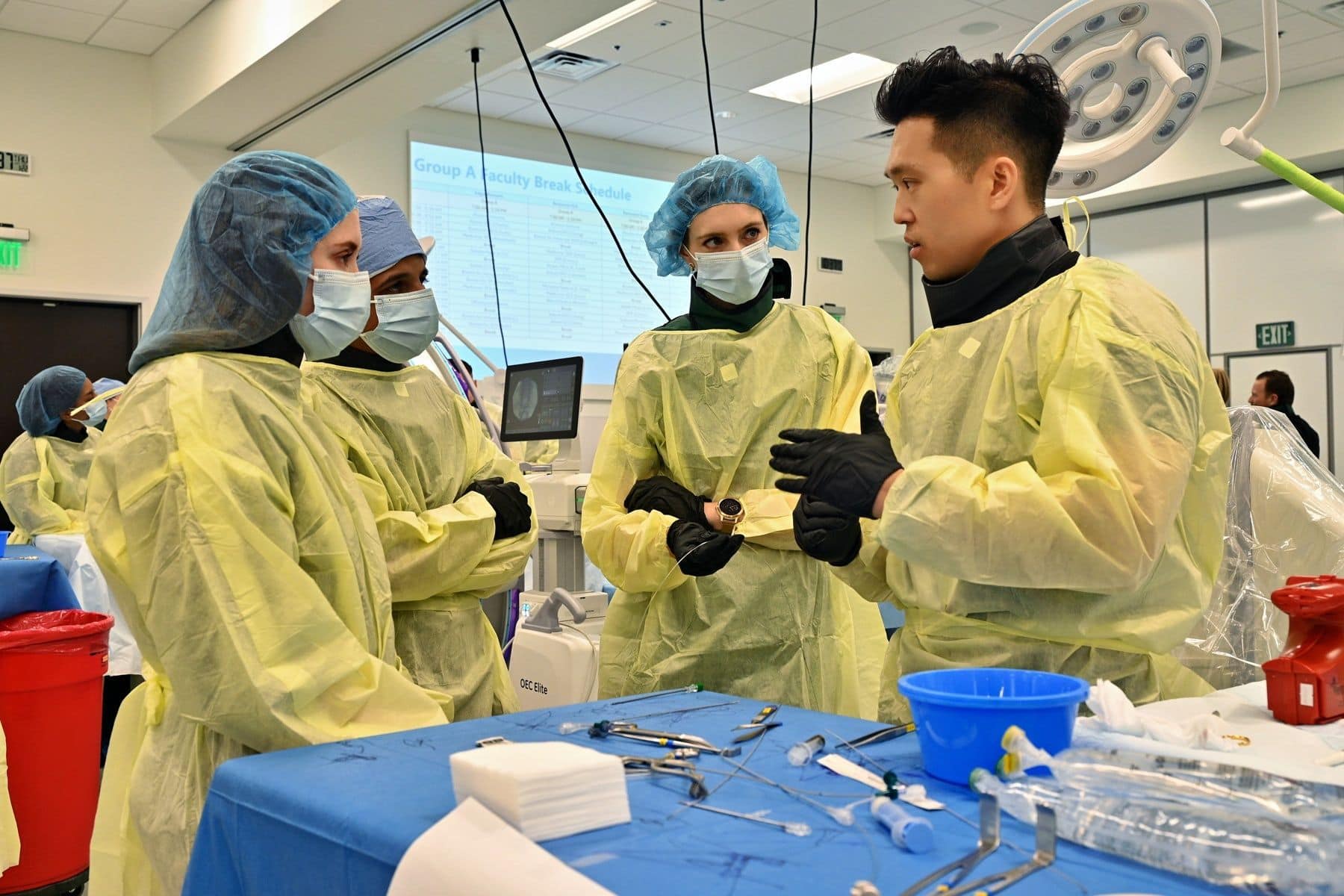Four medical professionals in yellow gowns, masks, and gloves converse in a brightly lit operating room with surgical instruments and equipment around them, as a presentation is projected on a screen in the background.