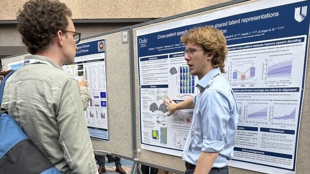 Two people discuss a scientific poster at a conference. One gestures toward data and graphs on the poster, while the other listens attentively. The setting appears to be indoors with other posters nearby.