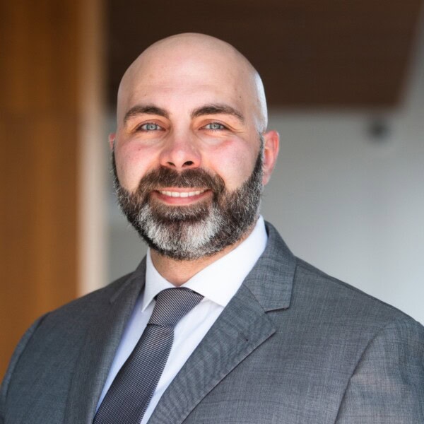 A man with a bald head and a full beard, wearing a gray suit, white shirt, and dark tie, smiles at the camera against a blurred indoor background.