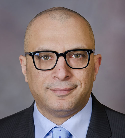 A man with glasses, short hair, and a suit and tie poses against a blurred gray and purple background.