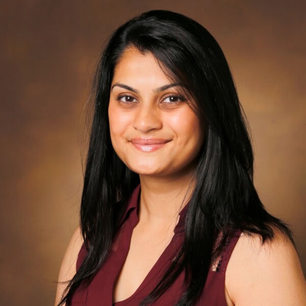 A woman with long dark hair smiles softly at the camera, wearing a sleeveless maroon top, in front of a plain brown background.