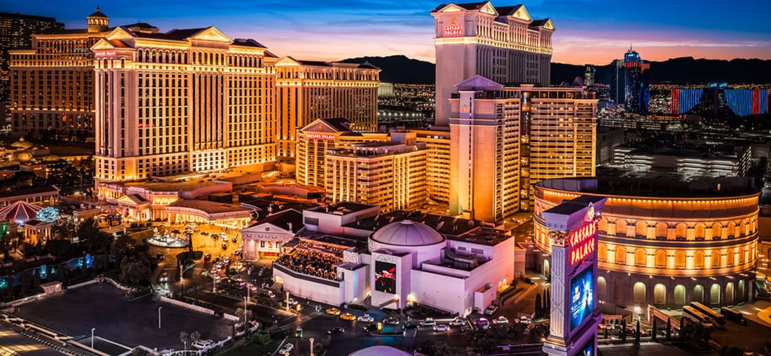 Las Vegas Strip at dusk featuring brightly lit hotels and casinos, including Caesars Palace, with vibrant lights and a colorful sunset in the background.