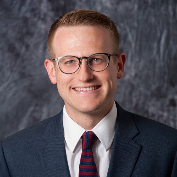 A man wearing glasses, a navy suit, a white shirt, and a red-and-blue striped tie smiles in front of a gray marbled backdrop.
