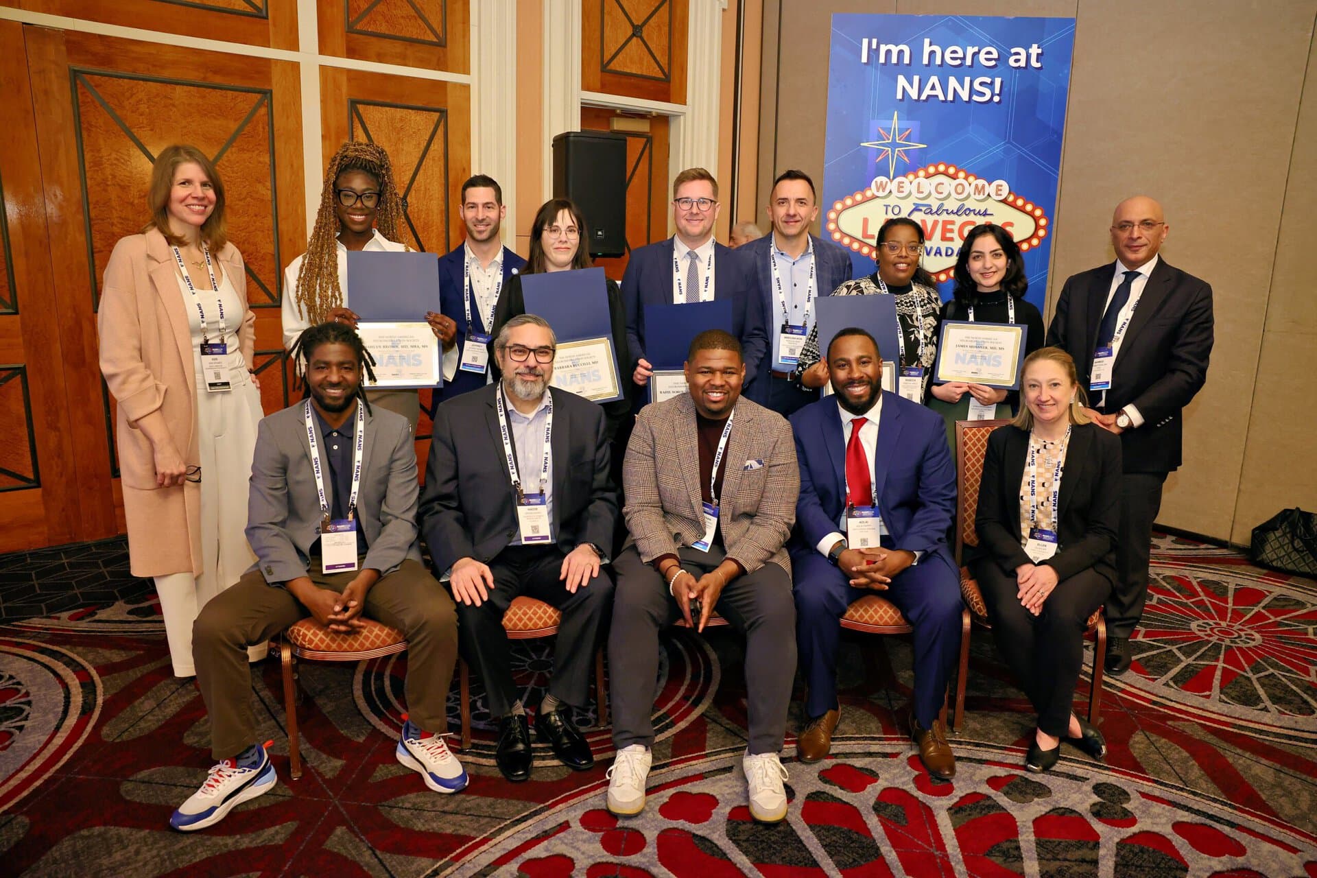 A group of people in business attire poses indoors, some holding certificates. They stand and sit in two rows, smiling at the camera in front of a Welcome to NANS sign on a patterned carpet.