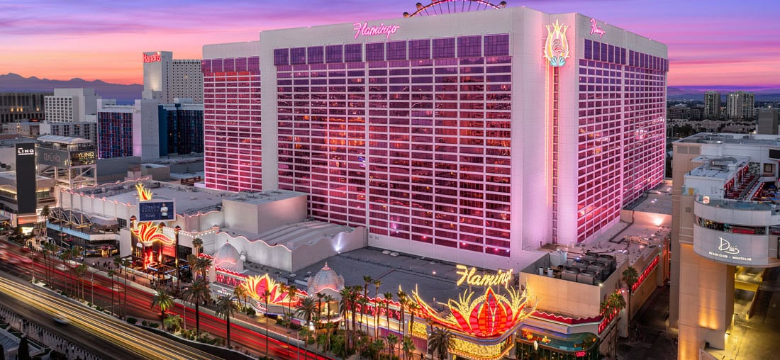 Aerial view of the Flamingo Las Vegas hotel and casino, glowing pink at sunset, with palm trees, neon lights, and surrounding cityscape.