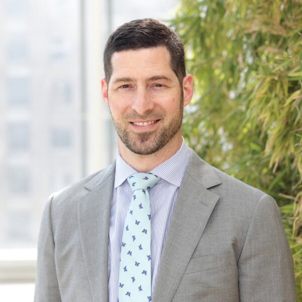 A man with short dark hair and a beard, wearing a light gray suit, blue dress shirt, and patterned tie, stands in front of a window and green plants.