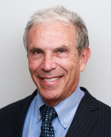 Smiling older man with gray hair wearing a dark pinstripe suit, light blue shirt, and patterned tie, posing in front of a plain light background.