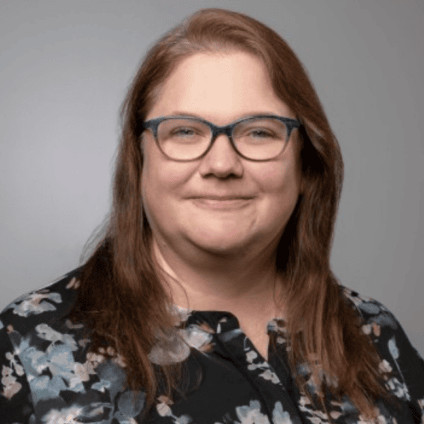 A woman with long brown hair and glasses smiles at the camera. She is wearing a black floral blouse and is posed in front of a plain gray background.