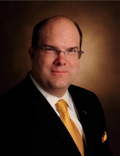 A man in a dark suit, white shirt, and yellow tie poses for a formal portrait against a brown gradient background. He has short hair, glasses, and a neutral expression.
