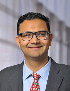 Smiling man wearing glasses, a gray suit, light blue shirt, and red patterned tie, posing in front of a blurred indoor background.