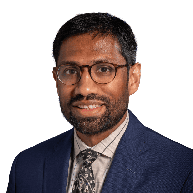 Man with short dark hair, glasses, and beard, wearing a blue suit jacket, patterned tie, and striped shirt, smiling at the camera against a plain white background.