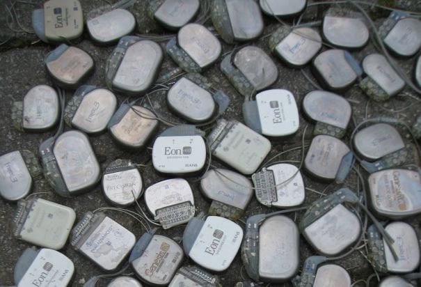 A pile of used pacemakers with visible wires and labels lies scattered on a rough, gray surface. Most devices are labeled Eon, and some serial numbers and inscriptions are visible on the metal casings.