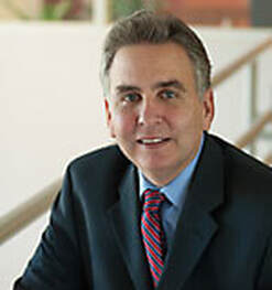 A man in a dark suit, blue shirt, and red-striped tie is smiling at the camera. He has short, grayish hair and is in an indoor setting with a blurred background.