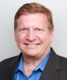 Smiling middle-aged man with short light brown hair, wearing a blue collared shirt and a dark blazer, pictured against a plain light background.