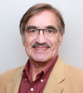 Smiling older man with light brown hair, glasses, and a mustache, wearing a tan blazer over a red striped shirt, against a plain light background.