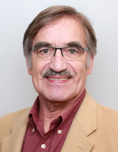 Smiling older man with gray hair, glasses, and a mustache, wearing a tan blazer over a maroon shirt, posed in front of a plain light background.