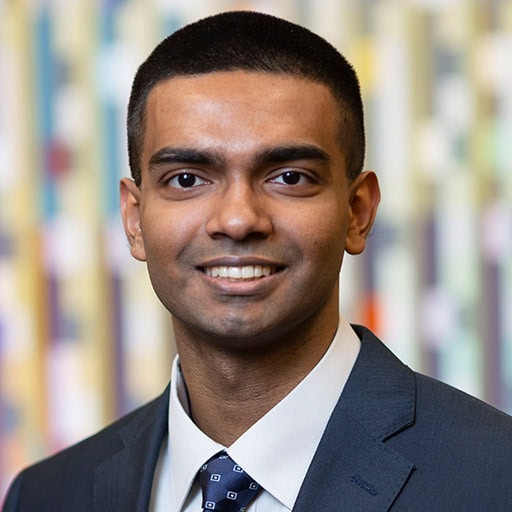 A man in a suit and tie smiles at the camera. The background is blurred with various vertical stripes of color.