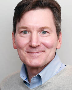 A man with short brown hair and blue eyes smiles at the camera. He is wearing a light blue collared shirt under a beige sweater, with a neutral gray background.