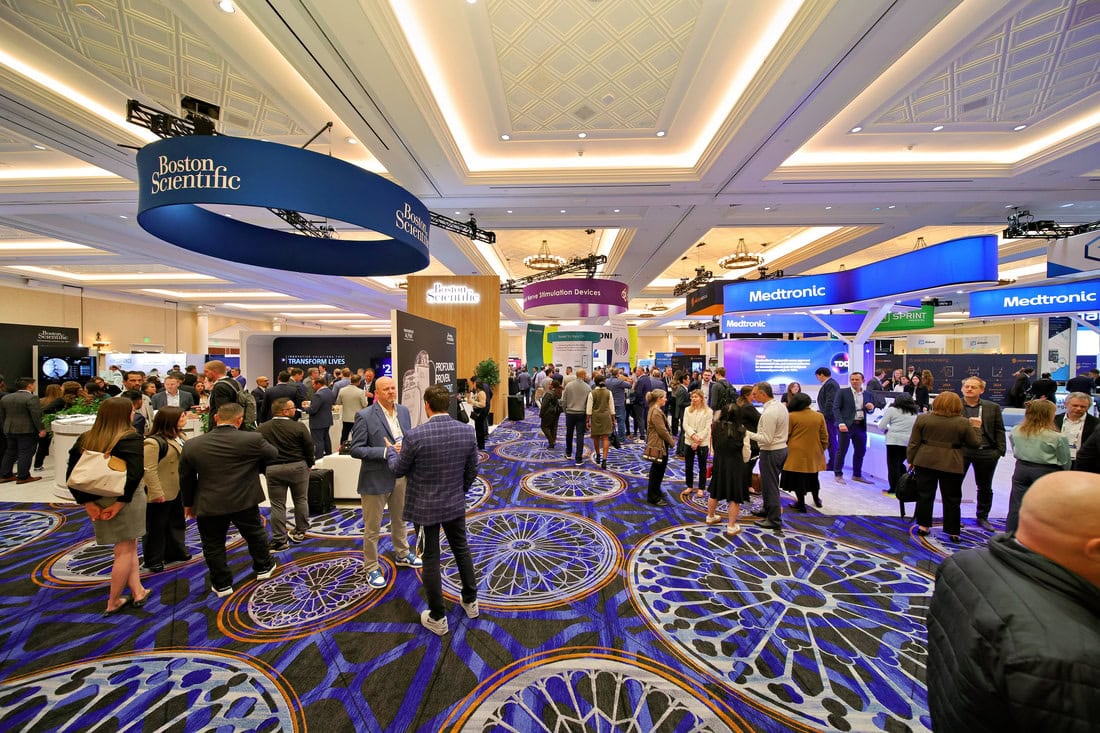 Large convention hall with groups of people networking near exhibitor booths, including Boston Scientific and Medtronic. The space features patterned blue carpeting and bright, modern lighting.