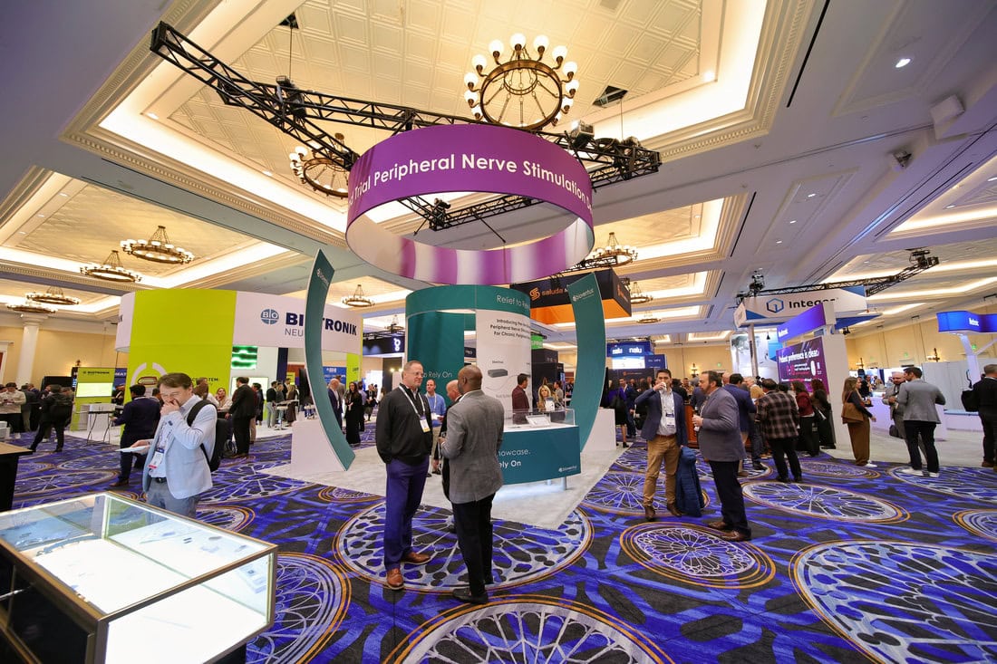 A busy medical conference with people networking among booths, including a prominent Peripheral Nerve Stimulation display, under elegant chandeliers in a large, carpeted hall.