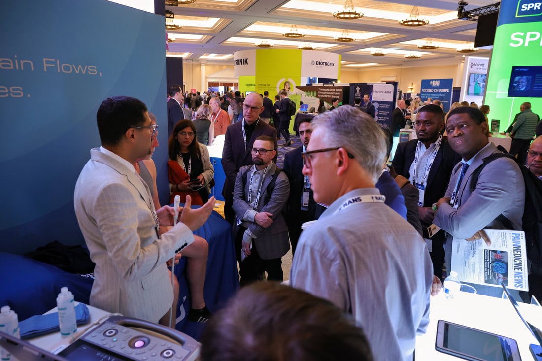 A man in a light suit speaks to a group at a technology conference booth. Attendees listen attentively, some holding conference badges and notepads, with bright screens and banners in the background.