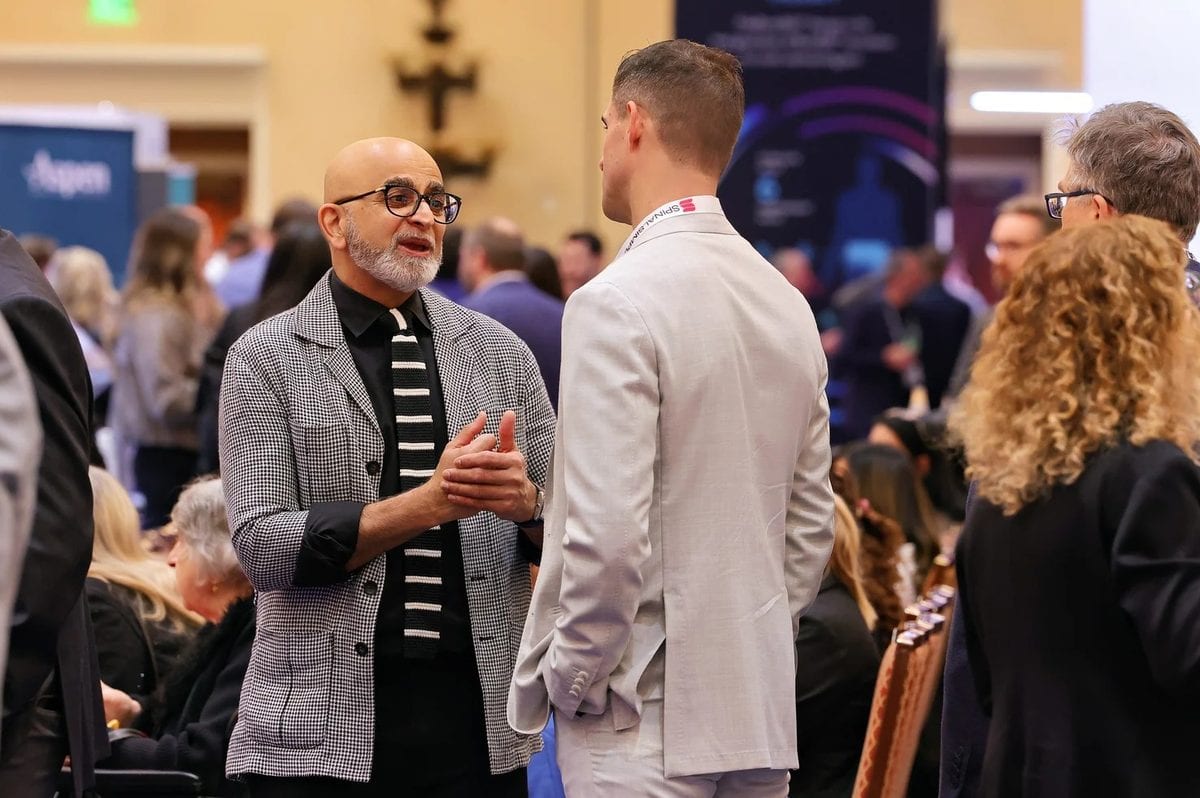 a man with glasses and a beard wearing a houndstooth blazer speaks to another man in a light colored suit during a conference.