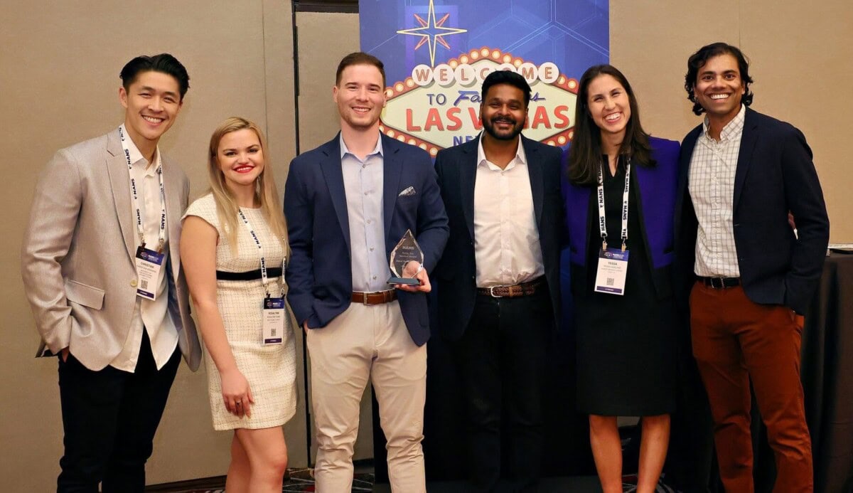 six people dressed in business attire stand together and smile at an event. one person in the center holds a glass award. a las vegas themed welcome sign is in the background.
