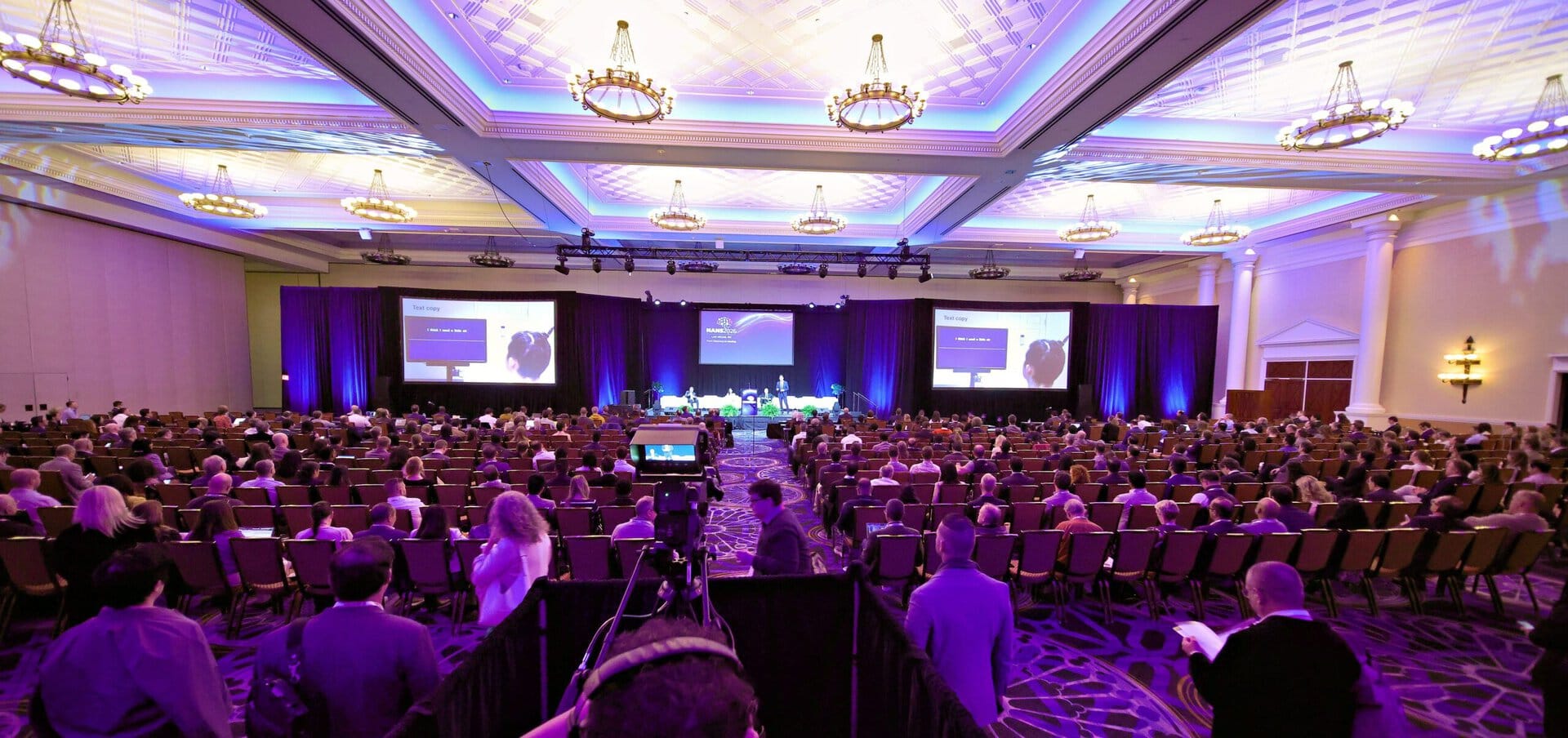 A large audience sits in a conference hall facing a stage with multiple speakers and presentation screens under elegant chandeliers; purple lighting highlights the room.